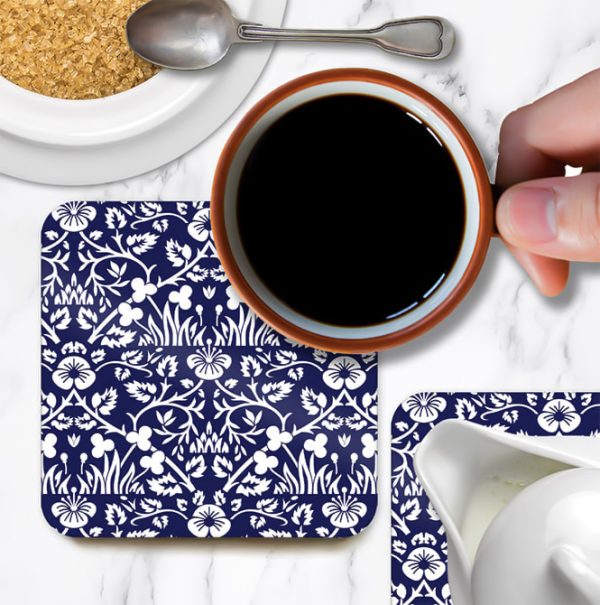 A blue coaster on table and coffee cup. Coaster is named Eyebright by William Morris.